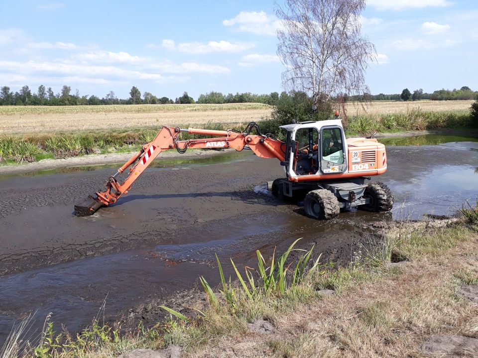 Mobilbagger räumt einen Teich, im Hintergrund Felder und Bäume