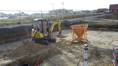 Minibagger hebt ein Fundament aus, daneben ein Betonkübel